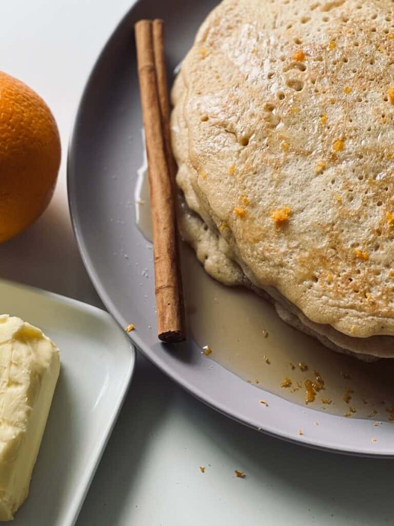 Stack of pancakes on a plate, topped with maple syrup and orange zest, decorated with a cinnamon stick, an orange and block of butter.
