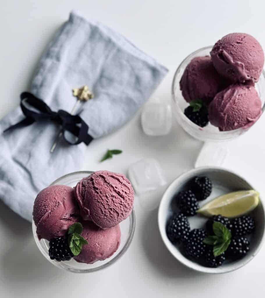 Two glass cups filled with homemade blackberry and lemon ice cream and a small bowl full of fresh blackberries and a lemon wedge.