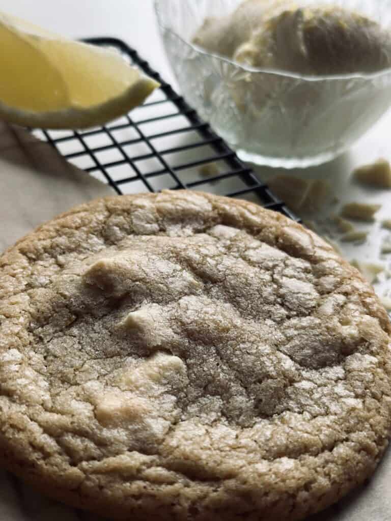 White chocolate and lemon cookie on a wire cooling rack, next to a bowl of ice cream and a wedge of lemon.