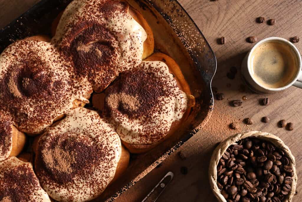 Tiramisu cinnamon rolls with a cup of espresso and a pot of roasted coffee beans.